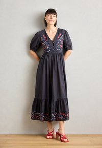Dunkelblaues, knielanges Kleid mit Puffärmeln, V-Ausschnitt und buntem Blumenstickerei. Ausgestellter Rock mit rüschiertem Saum. Kombiniert mit roten Sandalen.