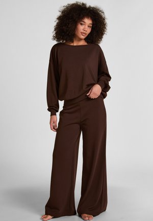 Vrouw met krullend haar, gekleed in een losse bruine top met lange mouwen en brede broekspijpen, staande op blote voeten tegen een effen grijze achtergrond.