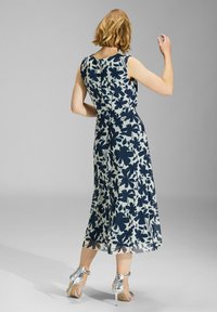 Kwiatowa sukienka midi z granatowym wzorem na jasnym tle, z bezrękawowym fasonem i przezroczystym dołem, zestawiona z srebrnymi szpilkami.