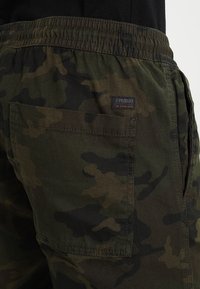 Maskáčové cargo kraťasy v tmavě zelené barvě se strukturovanou tkaninou, s elastickým pasem a vyšitým logem na zadní kapse.