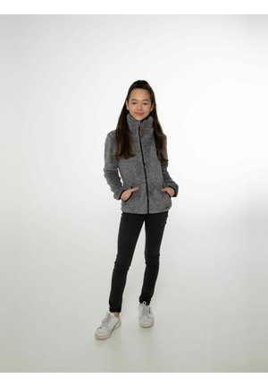 Chaqueta de forro polar gris con cuello alto, cierre de cremallera y dos bolsillos frontales. Combinada con jeans ajustados negros y zapatillas blancas.