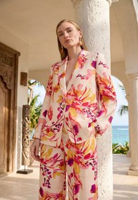 Vrouw in een blazer en broek met bloemenpatroon in roze, oranje en paars staat bij een wit stenen zuil met tropische planten en de oceaan op de achtergrond.