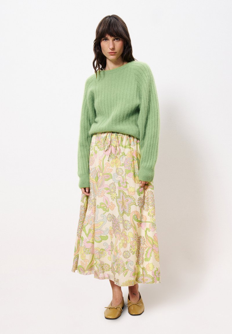 Grüner gerippter Strickpullover, maximaler Rock mit floralen Mustern in Pastellfarben und goldenen flachen Schuhen mit Schleifen. Lässige Passform und weiche Texturen.