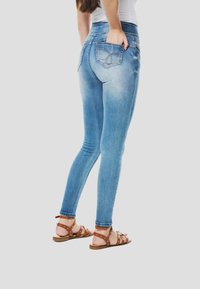 Strakke jeans in lichtblauw denim met een vervaagde wassing, hoge taille, en achterzakken met decoratieve stiksels. Bruine leren sandalen met studs.