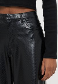 Calças pretas de imitação de couro com padrão trançado texturizado, apresentando bolsos frontais e cintura ajustada. O tecido tem um acabamento brilhante.