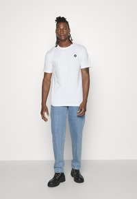 Vit bomullströja med korta ärmar, med en liten svart logotyp på bröstet, ihopkopplad med ljusblå denimjeans och svarta skor.