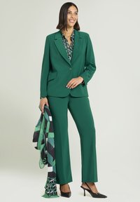 Grüner, maßgeschneiderter Anzug mit Sakko mit Reverskragen, gerade geschnittenen Hosen und einer gemusterten Bluse. Dazu High Heels und ein gemustertes Halstuch.
