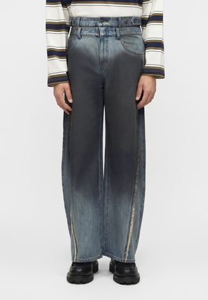 Τζιν με φαρδιά μπατζάκια και ξεβαμμένα σημεία, με πλαϊνές ραφές, φορεμένα με μαύρα chunky παπούτσια και ριγέ μακρυμάνικο πουκάμισο.