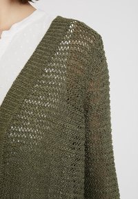 Cardigan lavorato a maglia verde oliva con un motivo a trama aperta, caratterizzato da una silhouette rilassata e un tessuto testurizzato per traspirabilità e comfort.