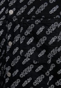 Svart tyg med ett upprepande vitt "CK50" logomönster. Tryckknappar och övre bröstficka med synliga sömnad detaljer.