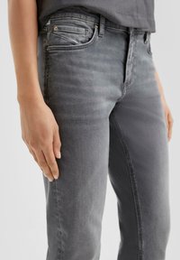 Vaqueros de mezclilla gris con un corte slim, que presentan un diseño de cinco bolsillos, costuras sutiles y una textura suave. Mano visible descansando sobre el muslo.