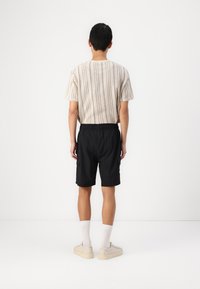 Pruhovaná pletená košeľa v odtieňoch hnedej, čierne šortky s elastickým pásom a svetlé tenisky. Jednoduché pozadie, zamerané na zadný pohľad na outfit.