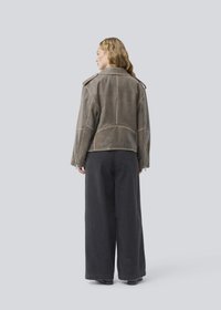 Brown leather jacket with contrasting stitching, wide collar, and zippers. Paired with loose black trousers. Rear view against a light background.