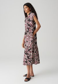 Abito maxi con maniche corte, base nera, ampi pattern di foglie bianche e bordeaux, gonna a strati, tasche laterali, abbinato a sandali neri.