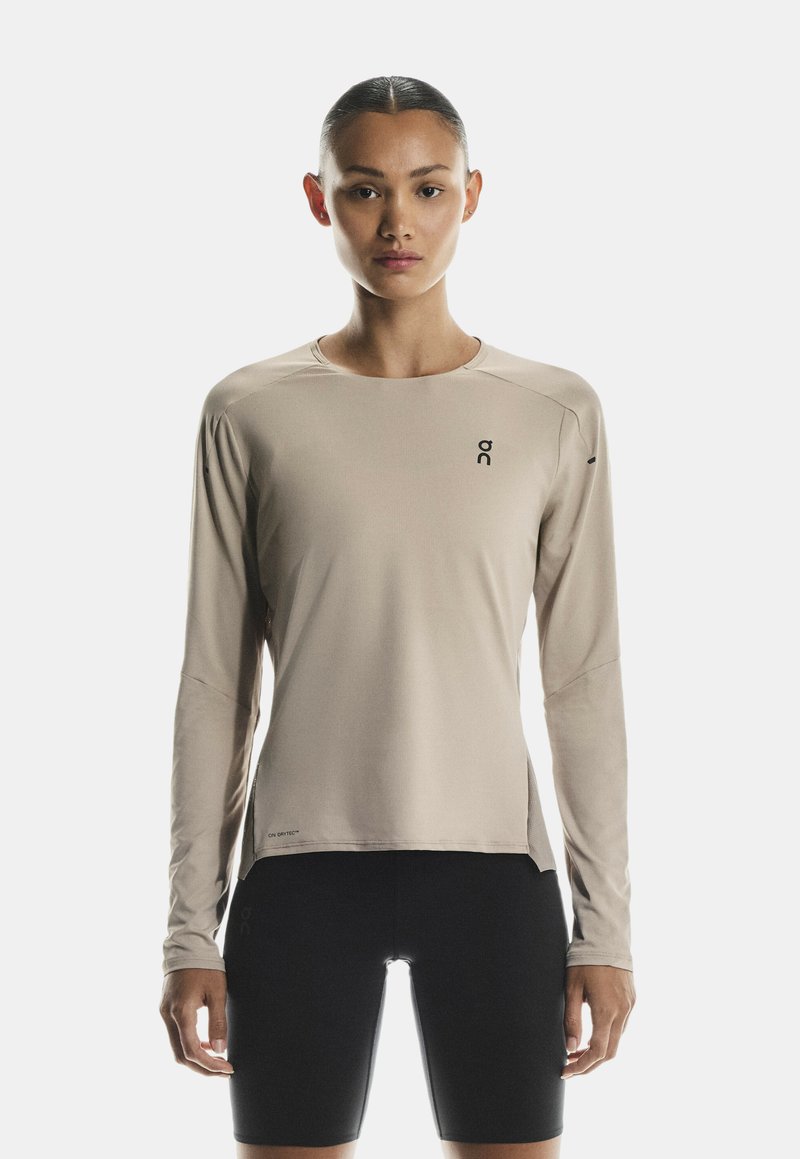 Langärmliges, beiges Sportshirt mit rundem Ausschnitt, seitlichen Schlitzen und Logo auf der Brust, kombiniert mit schwarzen Kompressionsshorts.