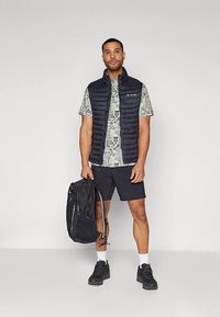 Muž stojí a má na sobě černou prošívanou vestu, vzorovanou košili s krátkým rukávem, černé šortky, bílé ponožky a černé tenisky. Drží černý batoh jednou rukou za popruh.