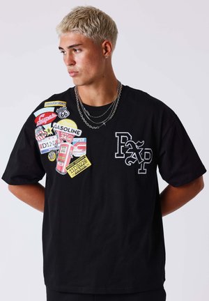 Giovane con capelli biondi corti che indossa una maglietta nera decorata con toppe colorate di stazioni di servizio e catene d'argento, che guarda verso sinistra.