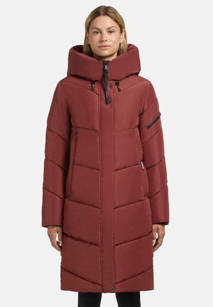 Femme portant un long manteau en duvet matelassé couleur rouille avec un col haut et des poches à fermeture éclair sur la poitrine et les manches, se tenant devant un fond uni.