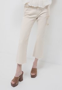 Pantaloni in denim beige con orlo grezzo, caratterizzati da un design slim. Abbinati a mules con piattaforma marroni, con punta aperta e accenti metallici.