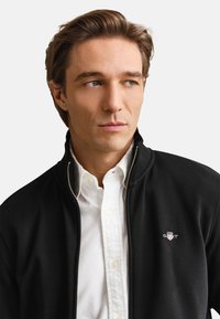 Młody mężczyzna o jasnobrązowych włosach, ubrany w białą koszulę na guziki i czarną kurtkę na suwak, lekko patrzy w lewo na jednolitym tle.