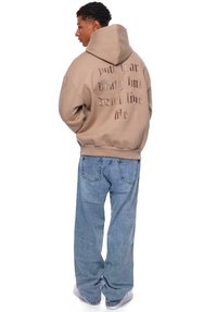 Sweatshirt beige à capuche avec un texte dans le dos, associé à un jean large bleu clair et des baskets blanches. Style décontracté, coupe relax.