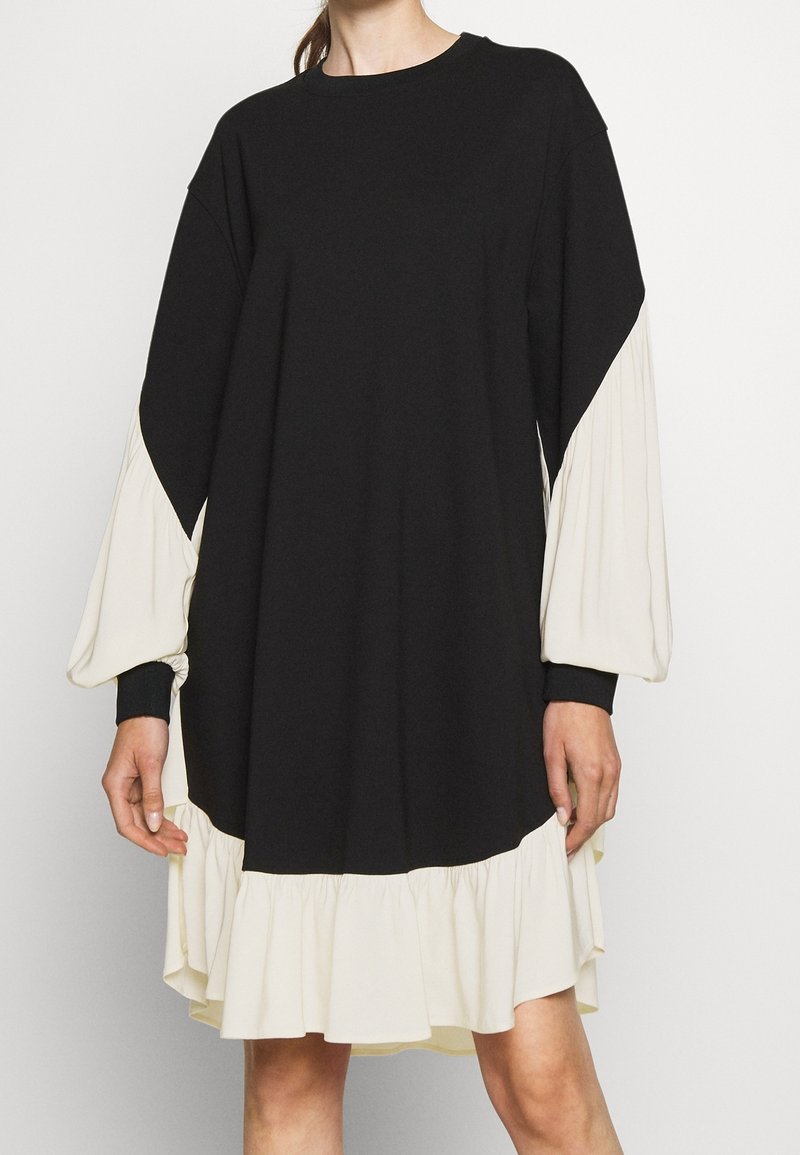 Robe noire à manches longues avec des manches ballon plissées crème et un ourlet à volants assorti, portée par une personne debout.