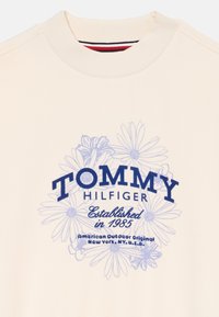 Μπλούζα κρεμ χρώματος με ριπ στο λαιμό. Διαθέτει μπλε λουλούδια γραφικών και το κείμενο "TOMMY HILFIGER". Οι εκτυπωμένες λεπτομέρειες περιλαμβάνουν το "Ιδρύθηκε το 1985".