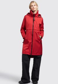 Chaqueta impermeable roja con cuello alto, cremallera frontal y capucha ajustable. Presenta dos bolsillos laterales y un corte holgado. Llevada sobre pantalones negros.