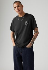 Sort bomulds t-shirt med et lille grafisk motiv på brystet, parret med mørk denim jeans. Korte ærmer og afslappet pasform.