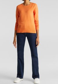 Oranger Strickpullover mit strukturierten horizontalen Streifen, rundem Halsausschnitt und langen Ärmeln. Kombiniert mit dunkelblauen Jeans und weißen Sneakern.