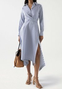 Vestido estilo cruzado a rayas en azul y blanco con cuello abotonado y mangas largas, con dobladillo asimétrico y aberturas. Bolso beige y tacones color cuero.