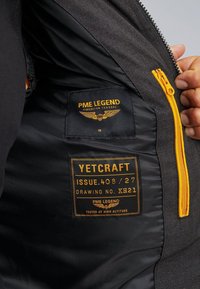 Čierny vnútorný kabát s oranžovým zipsom a štítkami, obsahujúci logo a informácie o produkte, hladká textúra a subtilný vzor podšívky.