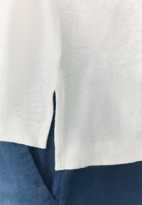 White cotton top with side slits, layered over dark blue fabric. The material appears soft, lightweight, and has a slightly textured finish.