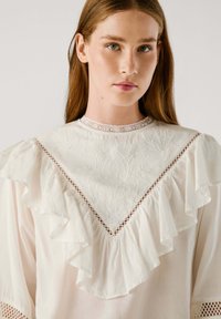 Blusa bianca con colletto a volant, pattern floreali ricamati e dettagli in pizzo. Il tessuto appare leggero e con una consistenza liscia.