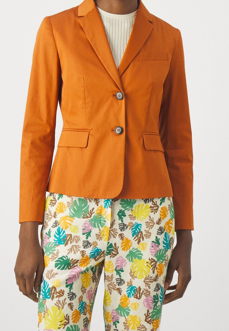 Personne portant un blazer orange sur un haut blanc côtelé et un pantalon coloré à motif de feuilles vertes, jaunes, roses, bleues et brunes.