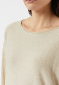 Beżowy dzianinowy sweter o gładkiej teksturze, szerokim dekolcie i subtelnym połysku. Charakteryzuje się luźnym krojem i prążkowanymi wykończeniami na krawędziach.