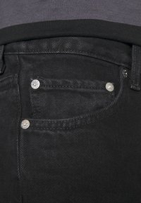 Gros plan d'une poche de jean en denim noir avec des rivets argentés et une ceinture sombre d'une chemise rentrée au-dessus.