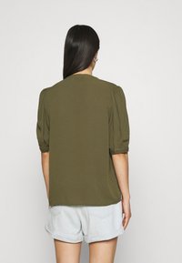 Blusa verde oliva con maniche corte e a sbuffo, materiale leggero, scollatura rotonda e vestibilità comoda, abbinata a pantaloncini di jeans chiari.