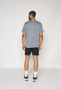 Grijze camouflage-patronen t-shirt met korte mouwen, gecombineerd met zwarte shorts. Zwart-witte sneakers, witte sokken zichtbaar.