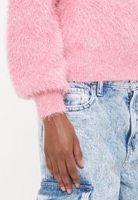 Fluffy roze trui met een ribbelrand en manchetten, gedragen over lichtblauwe spijkerbroek. De hand rust op de spijkerbroek en toont de textuur.