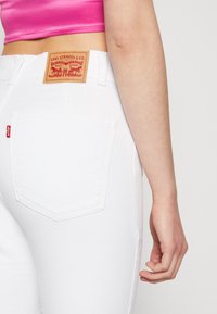 Los jeans de mezclilla blancos presentan un bolsillo trasero con un diseño de costura doble, un logo de etiqueta roja y un detalle de parche de cuero marrón de la marca.
