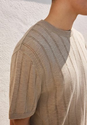 Person trägt einen beigen kurzärmeligen Strickpullover mit vertikalen offenen Strickstreifen und steht vor einer strukturierten weißen Wand.