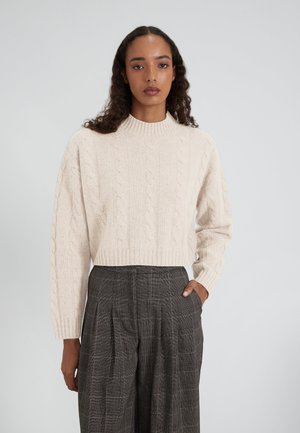Maglione a trecce spesse in morbido beige con collo alto, polsini a costine e vestibilità corta, abbinato a pantaloni grigi con motivo a quadri.