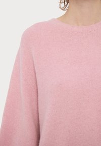 Απαλό ροζ πλεκτό πουλόβερ με στρογγυλή λαιμόκοψη που φοριέται από ένα άτομο, δείχνοντας το πάνω μέρος του κορμού και τη λεπτομέρεια του ώμου.