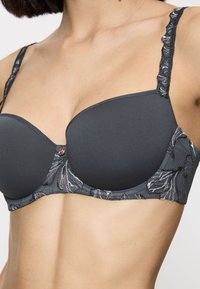 Nainen, joka pitää yllä mustaa ja tummanharmaata pehmeää rintaliiviä, jossa on kukkakuvioista pitsiä olan ja sivujen kohdalla, näkyy ylävartalo ja kaula.