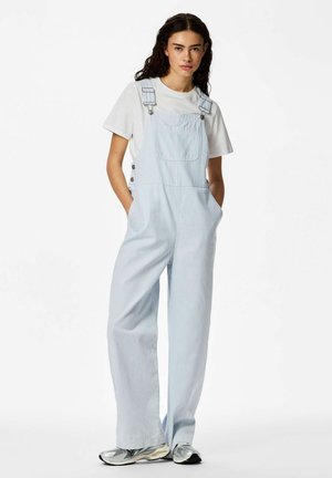 Ung kvinne iført lyseblåstripete overalls og hvit t-skjorte med hendene i lommene, stående i grå joggesko på hvit bakgrunn.