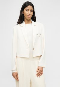 Γυναίκα που φοράει ένα μπεζ κοντό σακάκι με ένα κουμπί, παντελόνι καμπάνα που ταιριάζει και ένα ριμπ τοπ, όρθια μπροστά σε ένα απλό φόντο.