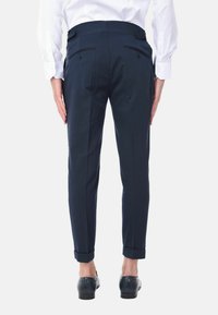 Pantaloni sartoriali blu navy con frontale piatto, due tasche posteriori e risvolti alle caviglie, abbinati a una camicia bianca a maniche lunghe e scarpe nere.