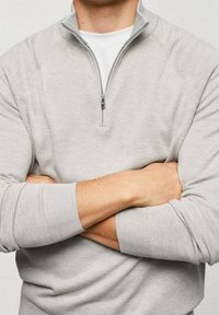 Maglione grigio chiaro con zip a un quarto e polsini a coste. Il tessuto appare morbido con una trama liscia. Indossato sopra una T-shirt bianca.