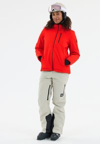 Rød isoleret jakke med en lynlåslomme; beige skibukser med sorte detaljer; sorte skistøvler; hvid hjelm og tonede goggles.